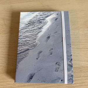 Può includere: Un taccuino rettangolare con una copertina fotografica che ritrae la riva di una spiaggia sabbiosa con delicate onde oceaniche. Diverse impronte umane sono visibili sulla sabbia bagnata e un elastico bianco tiene il taccuino chiuso saldamente.
