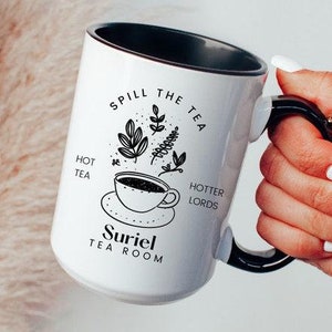 Pode incluir: Caneca de cer&acirc;mica branca com borda e al&ccedil;a pretas. A caneca tem um design preto e branco que diz "Spill the Tea" com uma x&iacute;cara de ch&aacute; e folhas. O texto "Hot Tea" e "Hotter Lords" est&atilde;o de cada lado da x&iacute;cara de ch&aacute;. O texto "Suriel Tea Room" est&aacute; na parte inferior da caneca.