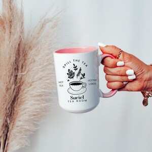 Pode incluir: Caneca de cer&acirc;mica branca com uma borda rosa e um design preto que diz "Spill the tea, hot tea, hotter lords, Suriel Tea Room". O design tamb&eacute;m inclui uma x&iacute;cara de ch&aacute; e um bule.