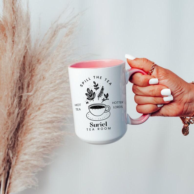 Pode incluir: Caneca de cer&acirc;mica branca com uma borda rosa e um design preto que diz "Spill the tea, hot tea, hotter lords, Suriel Tea Room". O design tamb&eacute;m inclui uma x&iacute;cara de ch&aacute; e um bule.