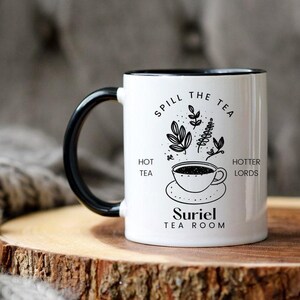 Pode incluir: Caneca de cer&acirc;mica branca com al&ccedil;a e borda pretas. A caneca tem um design preto e branco que diz "Spill the Tea" com uma x&iacute;cara de ch&aacute; e folhas. O texto tamb&eacute;m diz "Hot Tea" e "Hotter Lords". A parte inferior da caneca diz "Suriel Tea Room".
