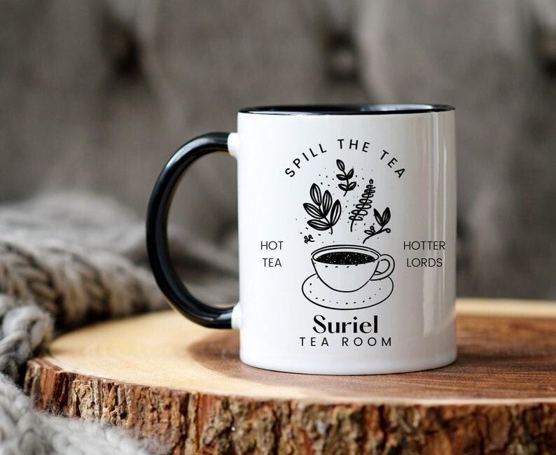 Pode incluir: Caneca de cer&acirc;mica branca com al&ccedil;a e borda pretas. A caneca tem um design preto e branco que diz "Spill the Tea" com uma x&iacute;cara de ch&aacute; e folhas. O texto tamb&eacute;m diz "Hot Tea" e "Hotter Lords". A parte inferior da caneca diz "Suriel Tea Room".