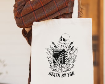 May include: A white tote bag with a black and white illustration of a skeleton holding a book. The text "DEATH BY TBR" is printed below the illustration.