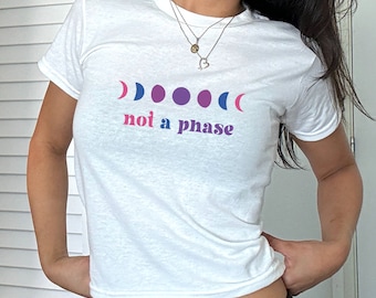 May include: White t-shirt with a graphic design featuring a series of moon phases in shades of pink, purple, and blue. The phrase "not a phase" is printed below the moon phases in pink and purple. The shirt has short sleeves.