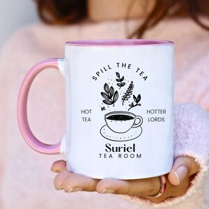 Pode incluir: Caneca de cer&acirc;mica branca com borda e al&ccedil;a rosa. A caneca tem um design preto que diz "Spill the Tea" com uma ilustra&ccedil;&atilde;o de uma x&iacute;cara de ch&aacute; e um bule. O texto "Hot Tea" e "Hotter Lords" est&aacute; em ambos os lados da ilustra&ccedil;&atilde;o. O texto "Suriel Tea Room" est&aacute; na parte inferior da caneca.