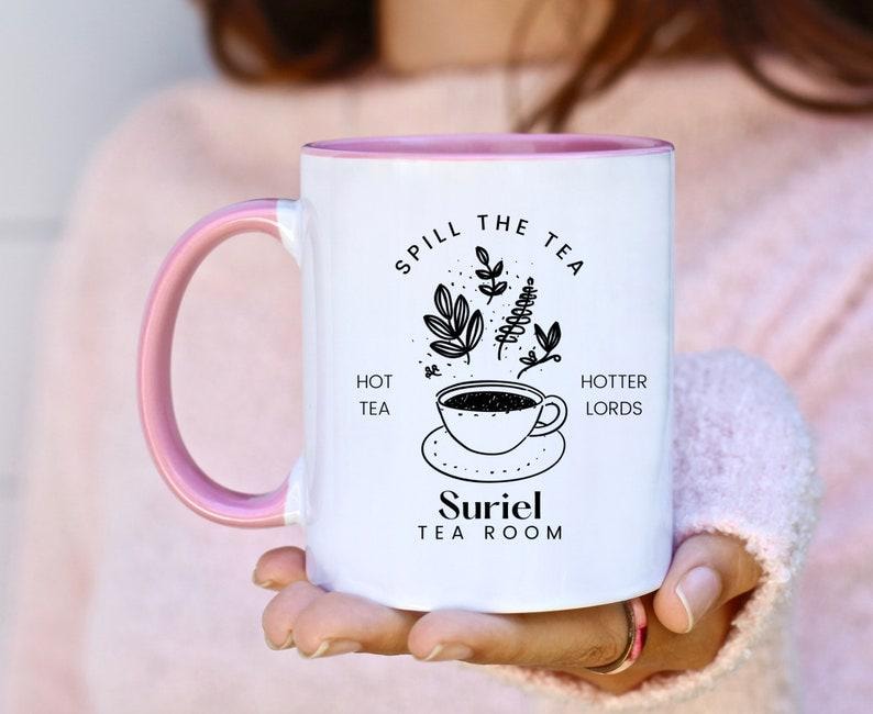 Pode incluir: Caneca de cer&acirc;mica branca com borda e al&ccedil;a rosa. A caneca tem um design preto que diz "Spill the Tea" com uma ilustra&ccedil;&atilde;o de uma x&iacute;cara de ch&aacute; e um bule. O texto "Hot Tea" e "Hotter Lords" est&aacute; em ambos os lados da ilustra&ccedil;&atilde;o. O texto "Suriel Tea Room" est&aacute; na parte inferior da caneca.