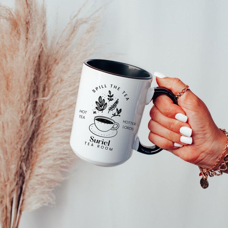 Pode incluir: Caneca de cer&acirc;mica branca com borda e al&ccedil;a pretas. A caneca tem um design preto e branco que diz "Spill the Tea" com uma x&iacute;cara de ch&aacute; e folhas. O texto "Hot Tea" e "Hotter Lords" est&atilde;o de cada lado da x&iacute;cara de ch&aacute;. O texto "Suriel Tea Room" est&aacute; na parte inferior da caneca.