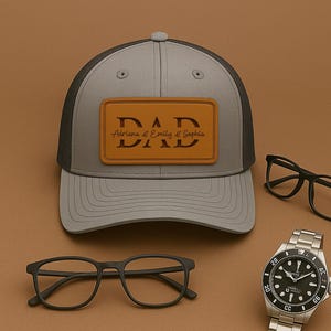 Może przedstawiać: Szara i czarna czapka z daszkiem z brązową skórzaną naszywką z napisem "DAD" i imionami "Adriana & Emily & Sophia". Ponadto dwie pary czarnych okularów i srebrny zegarek na rękę z czarną tarczą.