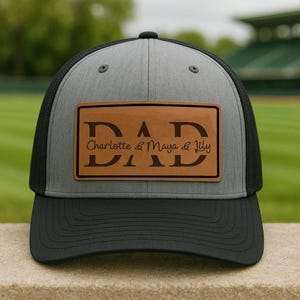 May include: A gray and black trucker hat with a black brim. The hat features a brown leather patch with the word "DAD" and the names "Charlotte & Maya & Lily" in a stylish font. The hat is likely for a father.