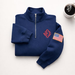 Könnte beinhalten: Marineblaues Sweatshirt mit Viertelreißverschluss, rotem Monogramm und einem amerikanischen Flaggenaufnäher auf dem Ärmel. Eine weiße Kaffeetasse befindet sich im Hintergrund. Das Sweatshirt hat einen silbernen Reißverschluss und gerippte Bündchen.