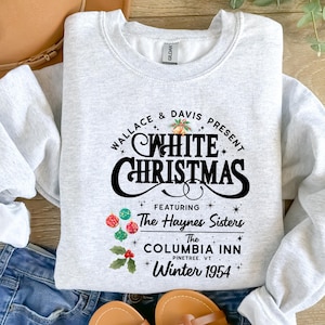 May include: A pale grey sweatshirt featuring a vintage-style design with the words "WHITE CHRISTMAS". The design includes the text "Wallace & Davis Present", "Featuring The Haynes Sisters", "The Columbia Inn Pinetree VT Winter 1954", and festive graphics.