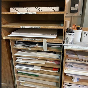 Op de afbeelding: Een houten rek met meerdere planken gevuld met stapels wit papier. Het papier is gelabeld met verschillende gewichten in gram, waaronder 170g, 250g en de woorden "Blanc Naturel" en "Extra Blanc".