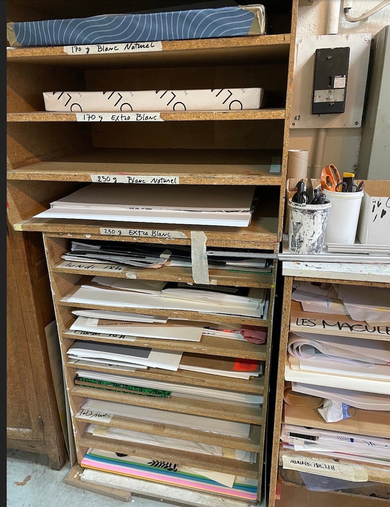 Op de afbeelding: Een houten rek met meerdere planken gevuld met stapels wit papier. Het papier is gelabeld met verschillende gewichten in gram, waaronder 170g, 250g en de woorden "Blanc Naturel" en "Extra Blanc".
