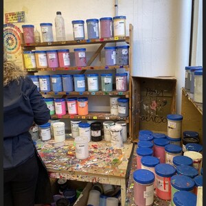Op de afbeelding: Een rommelige werkruimte met planken vol potten verf in verschillende kleuren. De potten zijn genummerd en hebben blauwe deksels. Er staan ook verschillende potten verf op een houten tafel, samen met een doos met het label "DECA" en een bord met de tekst "Kamer 9".