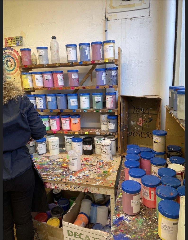 Op de afbeelding: Een rommelige werkruimte met planken vol potten verf in verschillende kleuren. De potten zijn genummerd en hebben blauwe deksels. Er staan ook verschillende potten verf op een houten tafel, samen met een doos met het label "DECA" en een bord met de tekst "Kamer 9".