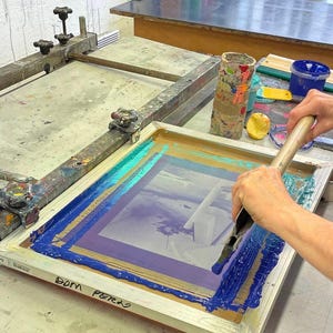 Op de afbeelding: Een persoon gebruikt een rakel om blauwe, turquoise en paarse verf over een zeefdrukframe te verspreiden. Het frame staat op een houten tafel met een bruin oppervlak. De tekst "DOM PERRO" staat op het frame.