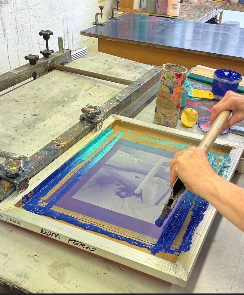Op de afbeelding: Een persoon gebruikt een rakel om blauwe, turquoise en paarse verf over een zeefdrukframe te verspreiden. Het frame staat op een houten tafel met een bruin oppervlak. De tekst "DOM PERRO" staat op het frame.