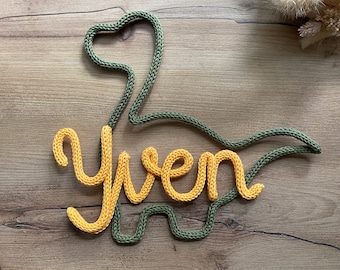 May include: A green knitted dinosaur outline with the name "Yven" spelled out in yellow yarn.