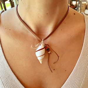 Könnte beinhalten: Eine braune Lederkordelkette mit einem weißen Muschelanhänger und bunten Perlen. Die Muschel hat braune Streifen.