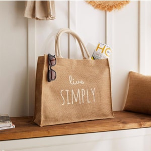 Puede incluir: Una bolsa de lona marr&oacute;n de yute con texto blanco que dice "live simply". La bolsa est&aacute; sentada sobre una superficie de madera y tiene un par de gafas de sol que sobresalen de la parte superior.