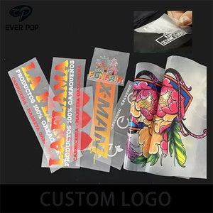 Könnte beinhalten: Nahaufnahme von mehreren transparenten Transferfolien aus Heißsiegelfolie mit verschiedenen aufgedruckten Designs. Die Designs beinhalten die Wörter "Funfair", "X-Man" und "La Flama" in bunten Lettern. Die Folien sind bereit, auf Stoff aufgebracht zu werden.
