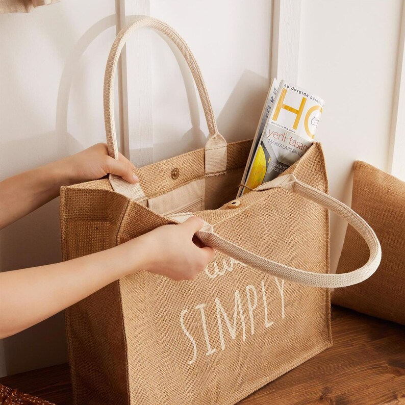 Pode incluir: Uma bolsa de juta marrom com al&ccedil;as brancas e o texto "and simply" impresso na frente. A bolsa est&aacute; cheia de revistas.
