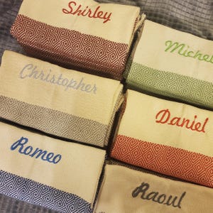 Puede incluir: Seis toallas personalizadas con diferentes nombres bordados. Las toallas son beige con un patrón de diamantes y una franja de color. Los nombres son Shirley, Michael, Christopher, Daniel, Romeo y Raoul.