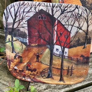 May include: A rectangular decorative tray featuring a rural landscape scene. The image depicts a red barn, trees, and figures in a field. The tray has rounded corners and a textured surface, suggesting a fabric or canvas material.