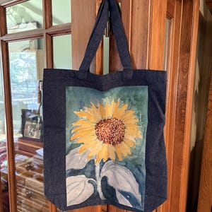 May include: A dark blue denim tote bag featuring a rectangular watercolor print of a large yellow sunflower with a brown center and green leaves. The bag has two matching denim shoulder straps and a small white tag on the side edge.