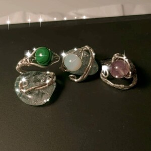 以下が含まれることがあります： 異なる色の石が付いた、銀色のワイヤーで巻かれた3つのリング。最初のリングには緑色の石、2番目のリングには白い石、3番目のリングには紫色の石がセットされています。