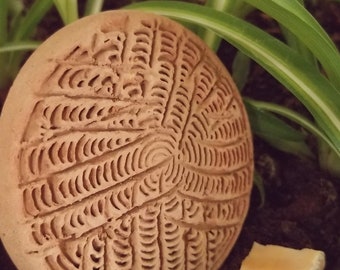 Puede incluir: Un disco redondo de cerámica de color terracota con un patrón detallado e intrincado de líneas y curvas. El disco está sentado en un lecho de tierra marrón.