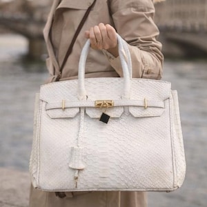 May include: A white handbag with a textured snakeskin pattern. The bag features gold-colored hardware, including a clasp, lock, and side details. The handles are white and the bag is held by a person wearing a tan trench coat.