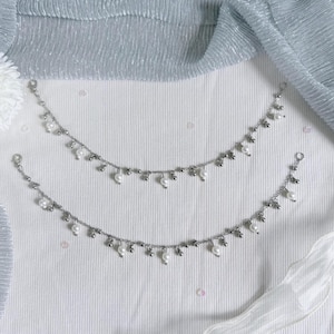 May include: Two silver-toned anklets featuring a delicate chain design adorned with small, evenly spaced white faux pearls and tiny metallic beads. The anklets are arranged in a curved, parallel formation, showcasing a consistent pattern of alternating beads.