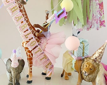 May include: A pink and gold birthday cake topper with a giraffe, elephant, and lion wearing party hats. The giraffe is holding a banner that says "Happy Birthday".