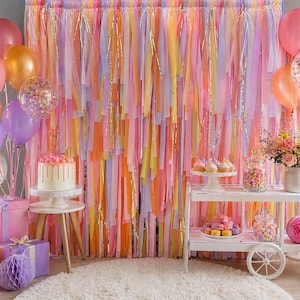 May include: A festive party scene with a colorful backdrop of hanging ribbons in shades of pink, orange, yellow, and gold. Balloons in various colors, a cake, and a cart filled with treats create a celebratory atmosphere. A white rug is on the floor.