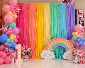 May include: A colorful party scene with a rainbow backdrop, balloons, and decorative elements. The scene includes a large rainbow arch, cloud cutouts, and a light-up number one, perfect for a birthday celebration. Balloons in various colors add to the festive atmosphere.