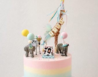 May include: A pastel rainbow-layered cake decorated with toy animal figurines, including a giraffe, zebra, elephant, and rhino. The giraffe has a party hat and a banner that says "YARA". The cake is on a white cake stand and has colorful sprinkles.