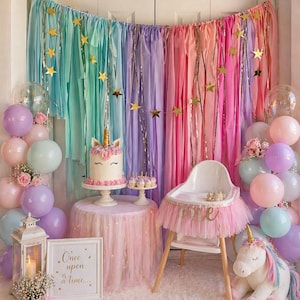 May include: A whimsical party scene with a unicorn theme. A cake, cupcakes, and a high chair are decorated with unicorn details. Pastel balloons, a backdrop of colorful streamers, and a plush unicorn complete the festive decor.
