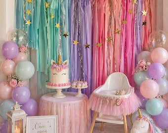 May include: A whimsical party scene with a unicorn theme. A cake, cupcakes, and a high chair are decorated with unicorn details. Pastel balloons, a backdrop of colorful streamers, and a plush unicorn complete the festive decor.