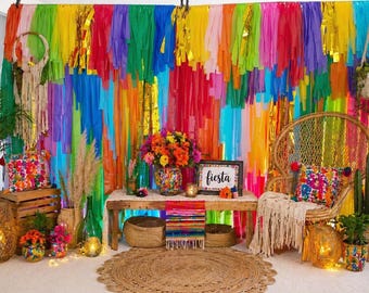 May include: A vibrant fiesta-themed display with a colorful backdrop of layered paper streamers. The scene includes a wooden bench, woven chair, and decorative pillows. A sign says "fiesta". Colorful flowers and plants add to the festive atmosphere.