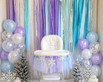 May include: A high chair decorated with a snowflake-themed tulle skirt, set against a backdrop of purple, blue, and silver streamers. Balloons and decorative trees complete the winter wonderland theme.