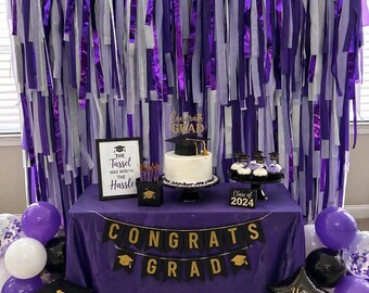 May include: A graduation party setup with a purple tablecloth, balloons, and decorations. A banner reads "Congrats Grad" with gold lettering. A cake and cupcakes are on the table, along with a sign that says "The Tassel Was Worth The Hassle."