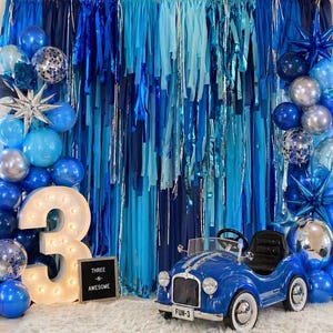 May include: A blue toy car with white tires and the text "FUN-3" sits in front of a backdrop of blue streamers and balloons. A large illuminated number "3" and a sign that reads "THREE -N- AWESOME" are also present, creating a festive birthday scene.