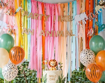 Op de afbeelding: Een feestelijke partyscène met een kleurrijke achtergrond van regenboog streamers en ballonnen. Een taart met een leeuwenfiguur en de woorden "Wild One" staat op een witte taartstandaard. De banner luidt "Party Like An Animal."