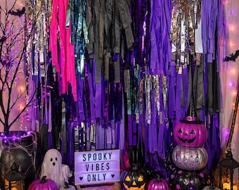 May include: A Halloween-themed display with a backdrop of purple, silver, black, and pink fringe. The scene includes pumpkins, a ghost, a sign that reads "Spooky Vibes Only," and decorative lights. A witch's hat sits atop a stack of pumpkins.