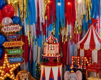 May include: A festive circus-themed party setup with a red, white, and blue color scheme. The scene includes a cake, a popcorn machine, balloons, and a sign that says "Welcome to the Carnival". A plush lion and elephant are also present.