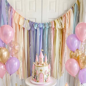 May include: A fairytale-themed party setup with a castle cake, balloons, and a decorative garland. The cake is white with pink accents, a castle design, and floral decorations. Balloons in pink, gold, and purple are arranged around the cake. A gold crown and star-shaped light add to the decor.