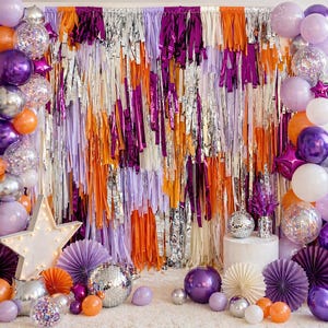 May include: A festive party scene with a backdrop of orange, purple, silver, and white fringe. Balloons in shades of purple, silver, and orange, along with disco balls and star-shaped lights, create a celebratory atmosphere. Paper fans and decorative elements complete the scene.