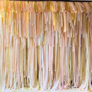 May include: A backdrop made of a variety of thin fabric strips in shades of pink, white, and gold. The strips are hanging from a horizontal rod and are arranged in a cascading pattern.