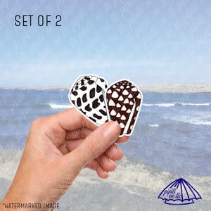 May include: Two seashell stickers, one with black spots on white, the other with brown spots on white, held in front of a beach scene. The text "SET OF 2" is at the top. The stickers are heart-shaped.